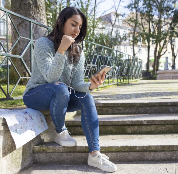 Serious pretty woman listening to music on city stairs parapet Serious pretty woman listening to music on city stairs parapet. Beautiful young lady sitting with paper map, smartphone and city view in background. Urban lifestyle concept.